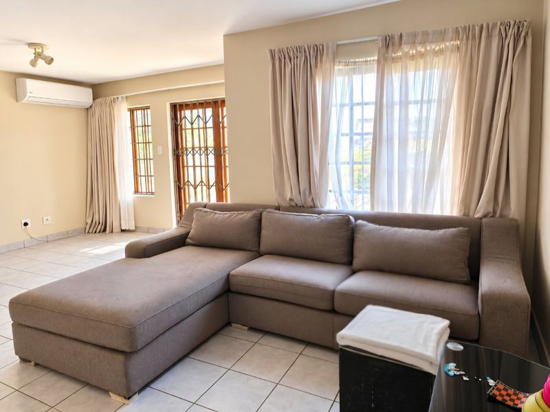 A bright and spacious living room featuring tiled flooring, a wall-mounted air conditioning unit, and secure access points with security gates and window bars.
