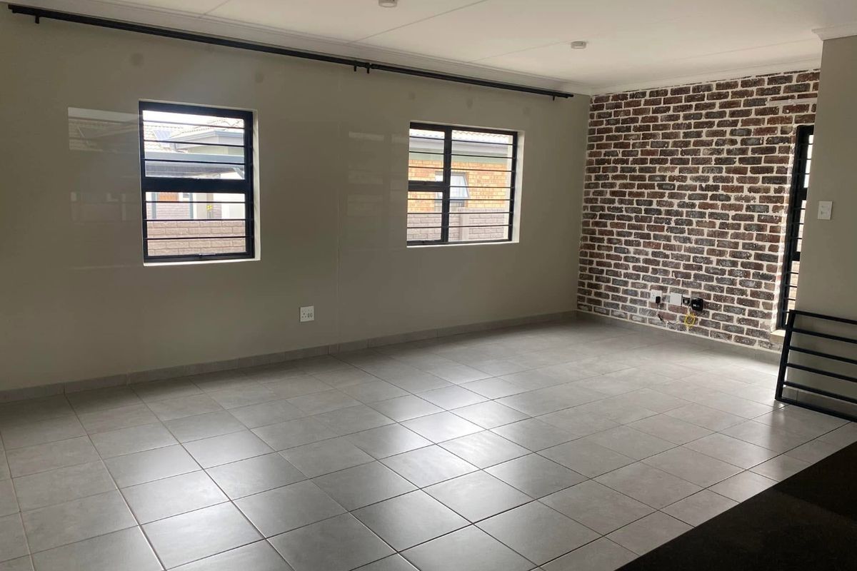Spacious living area featuring neutral ceramic floor tiles, an exposed brick feature wall, and large windows with security bars.