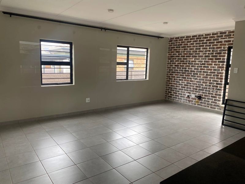 Spacious living area featuring neutral ceramic floor tiles, an exposed brick feature wall, and large windows with security bars.
