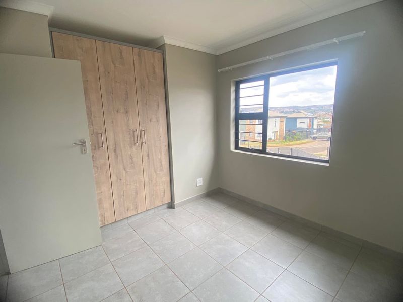 A bright, empty bedroom featuring modern light wood-finish built-in cupboards, neutral tiled flooring, and a large window with security bars.