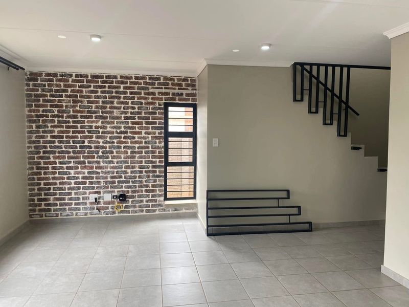 Spacious living area featuring an exposed brick accent wall, neutral tiled flooring, and a modern staircase with a black metal balustrade.