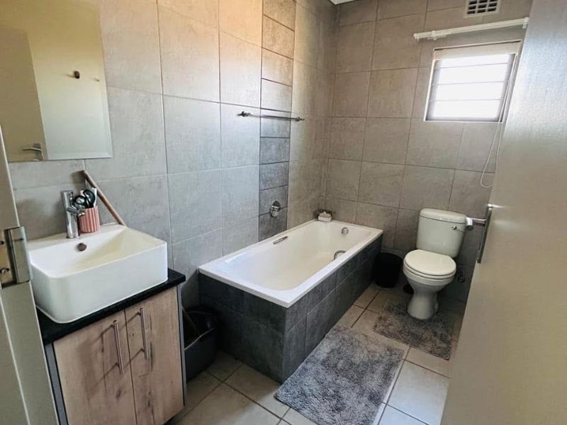 A modern bathroom featuring a built-in bathtub, a vanity cabinet with a basin, and a toilet, finished with neutral floor-to-ceiling wall tiles.