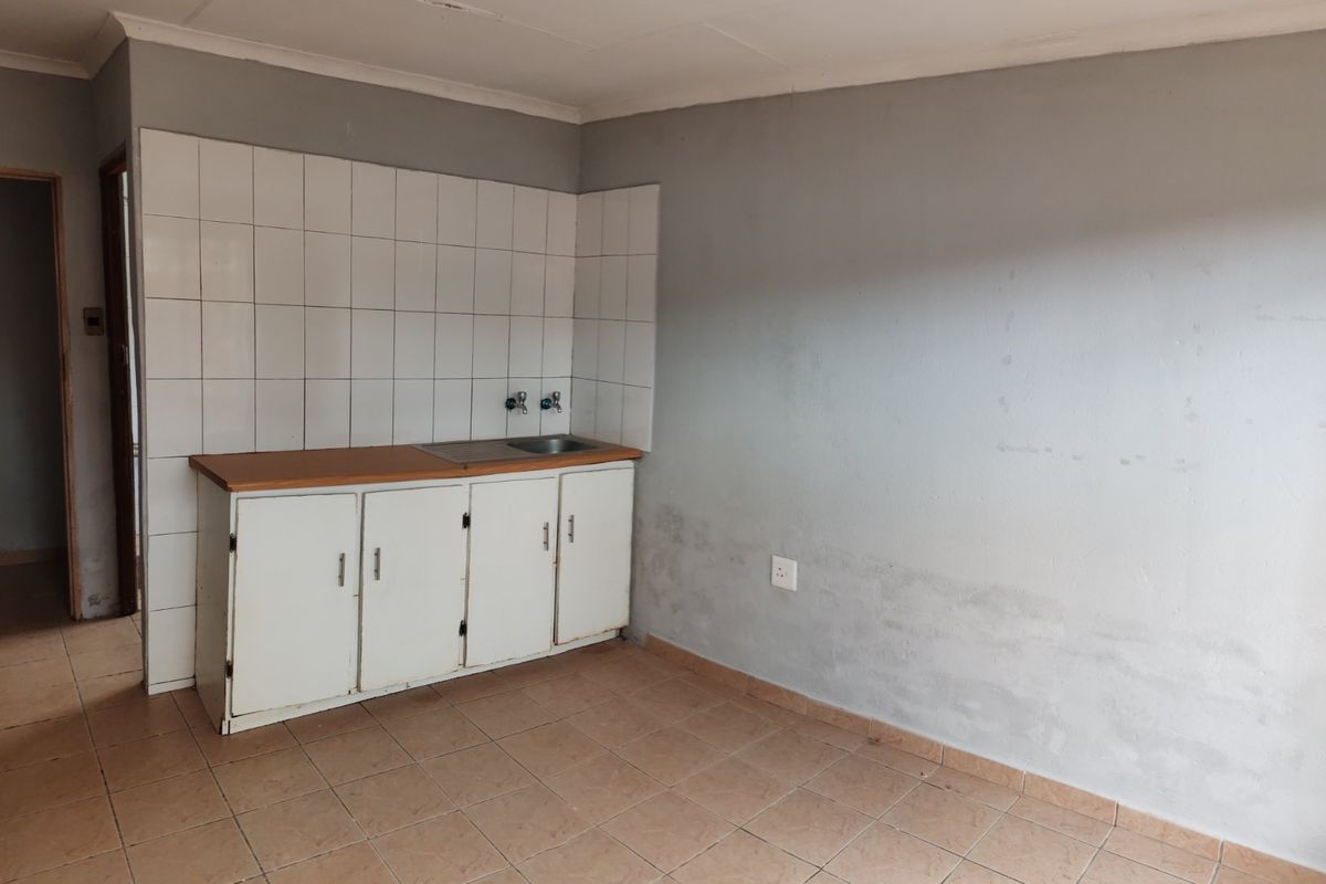 A compact, empty kitchen area featuring basic base cabinetry, a sink, tiled splashback, and tiled flooring, requiring cosmetic updates.