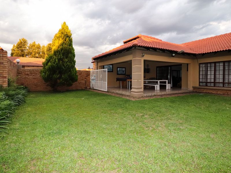 A rear view of the property featuring a well-maintained lawn, a covered patio area, and a brick boundary wall under a tiled roof.
