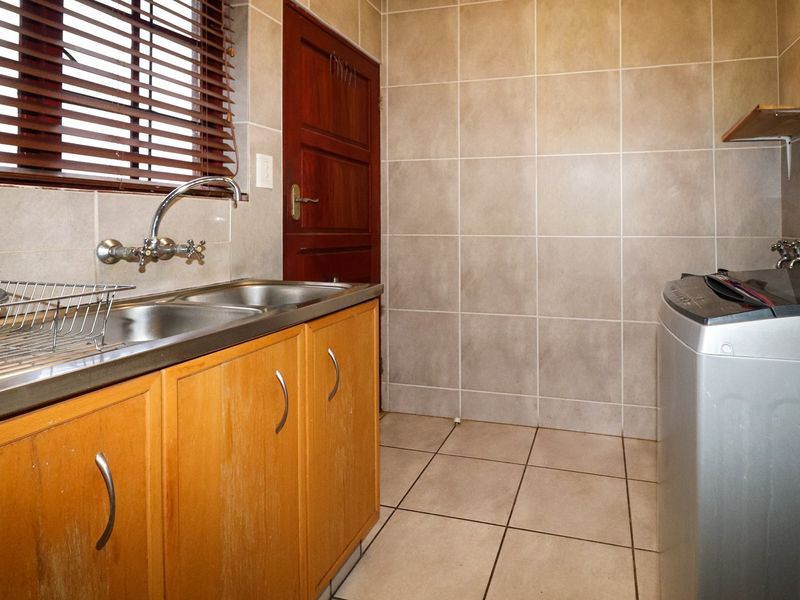 A functional laundry room featuring tiled walls and floors, built-in wooden cabinetry, a stainless steel double sink, and dedicated plumbing for a washing machine.