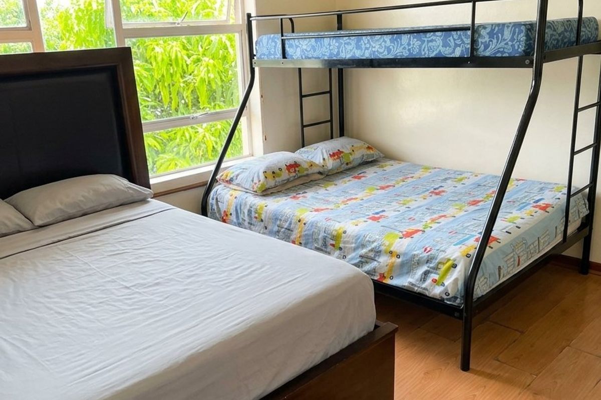 A functional bedroom featuring timber laminate flooring and large windows with venetian blinds, providing natural light and a view of the greenery outside.