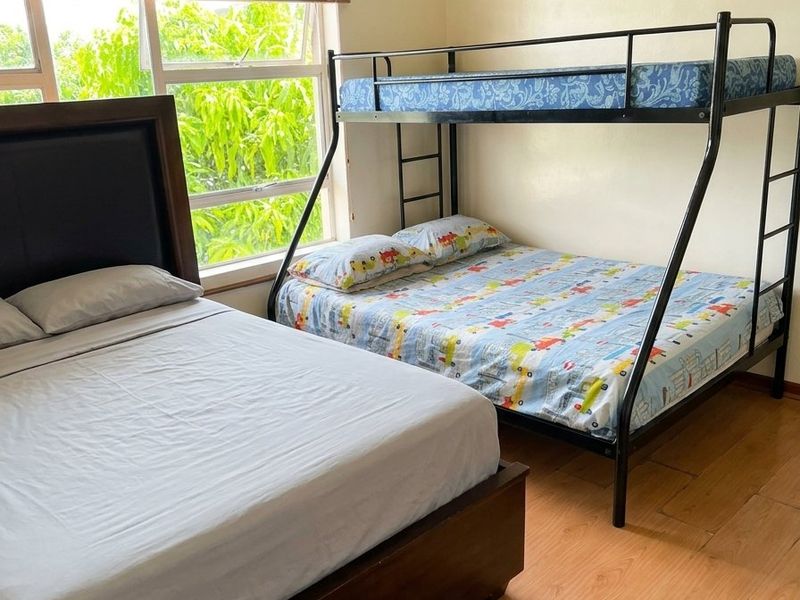 A functional bedroom featuring timber laminate flooring and large windows with venetian blinds, providing natural light and a view of the greenery outside.