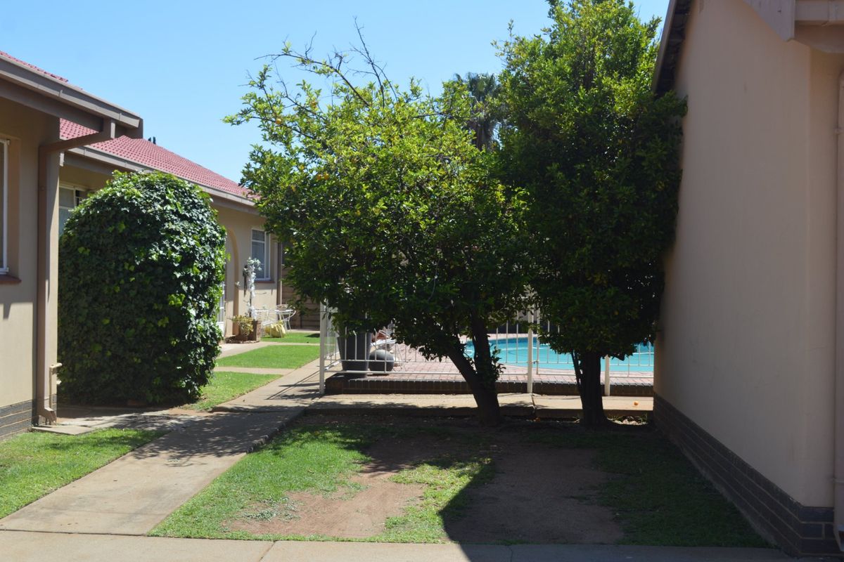 A rear exterior view of the apartment complex featuring a swimming pool, paved walkways, and established garden areas.
