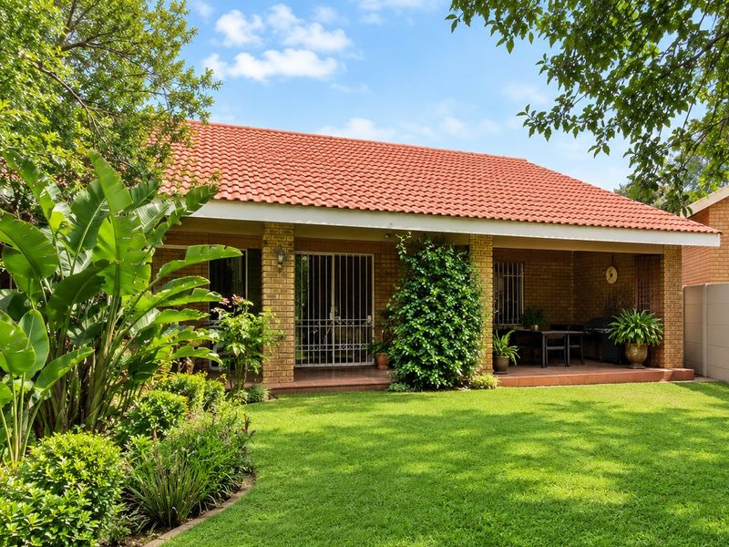A well-maintained rear exterior view featuring a covered patio, brick facade, tiled roof, and a lush, landscaped garden.