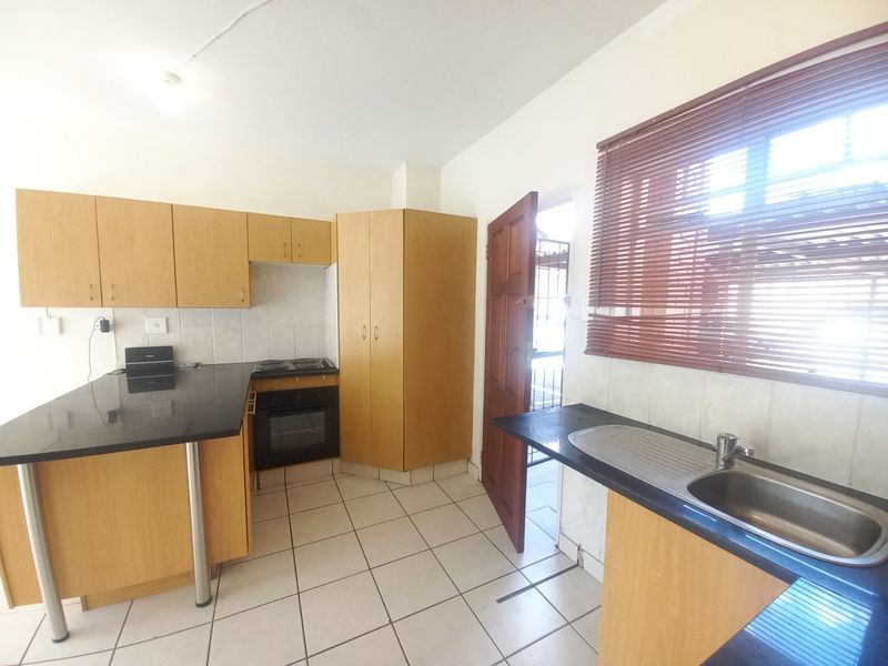 A functional kitchen featuring light wood cabinetry, dark laminate countertops, built-in oven and hob, and tiled flooring with direct access to an exterior door.
