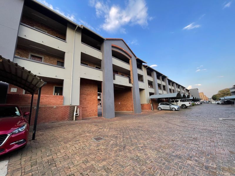 Exterior view of a multi-story apartment complex featuring brick and plaster facade, paved parking area, and covered carport structures.