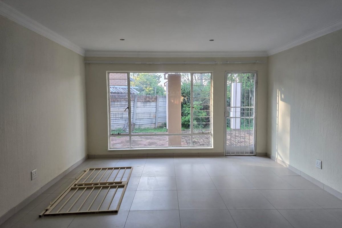A bright, empty living room featuring neutral tiled flooring, recessed lighting, and large windows providing natural light and access to the outdoor area.