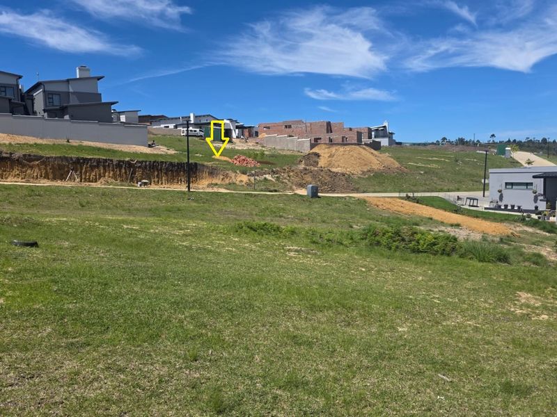 A vacant residential plot featuring a sloped landscape with an exposed earth bank, situated within a developing estate with surrounding modern homes.