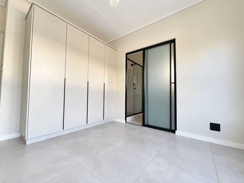 A modern, minimalist bedroom featuring neutral tiled flooring, extensive built-in cabinetry, and a sleek sliding glass door leading to an en-suite bathroom.