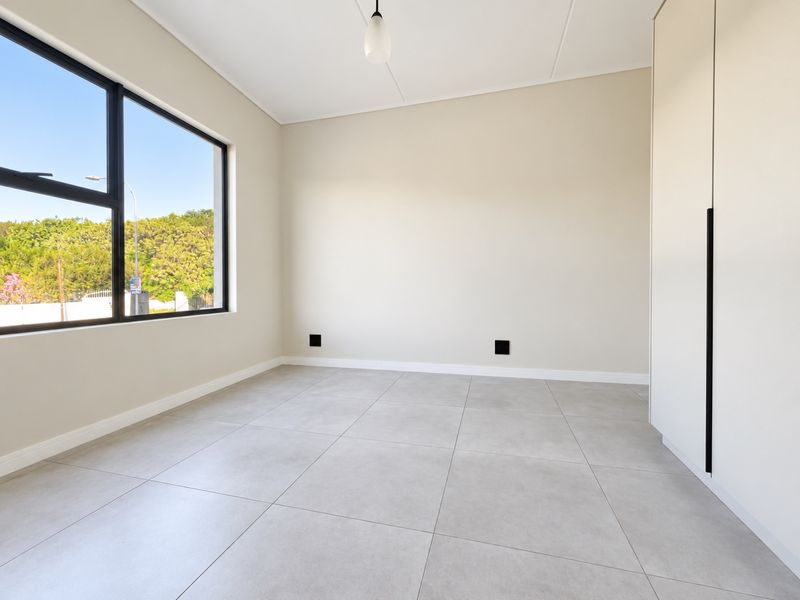 A bright, modern, and empty bedroom featuring neutral tiled flooring, built-in cupboards, and a large window providing ample natural light.