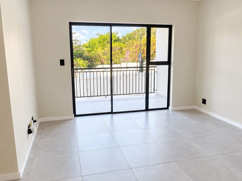A bright, modern, and empty living area featuring large format floor tiles, track lighting, and a black aluminium sliding door leading to a balcony.