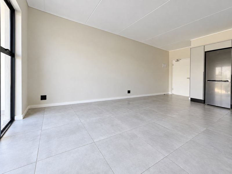 A bright, modern open-plan living area featuring neutral tiled flooring, clean finishes, and a large glass door providing ample natural light.