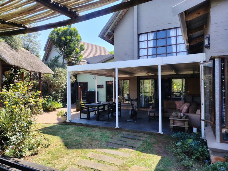 Spacious covered patio area featuring tiled flooring and a metal pergola, overlooking a landscaped garden with brick pathways and a thatched outdoor structure.