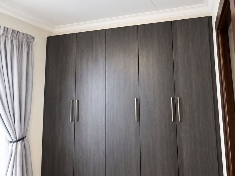 A clean bedroom interior featuring modern dark wood built-in cupboards, tiled flooring, and a ceiling light fixture.