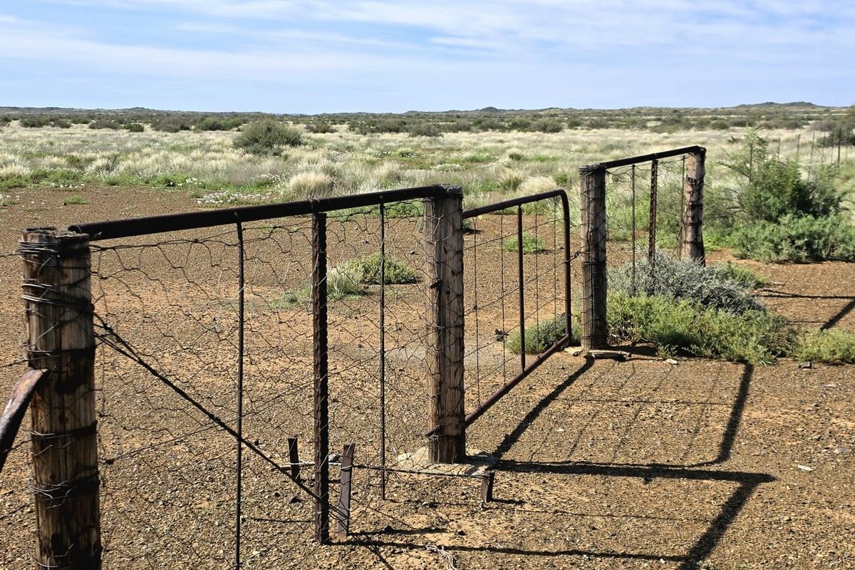 A view of a rural farm property featuring weathered timber fence posts, rusted wire mesh fencing, and a metal farm gate overlooking open veld.
