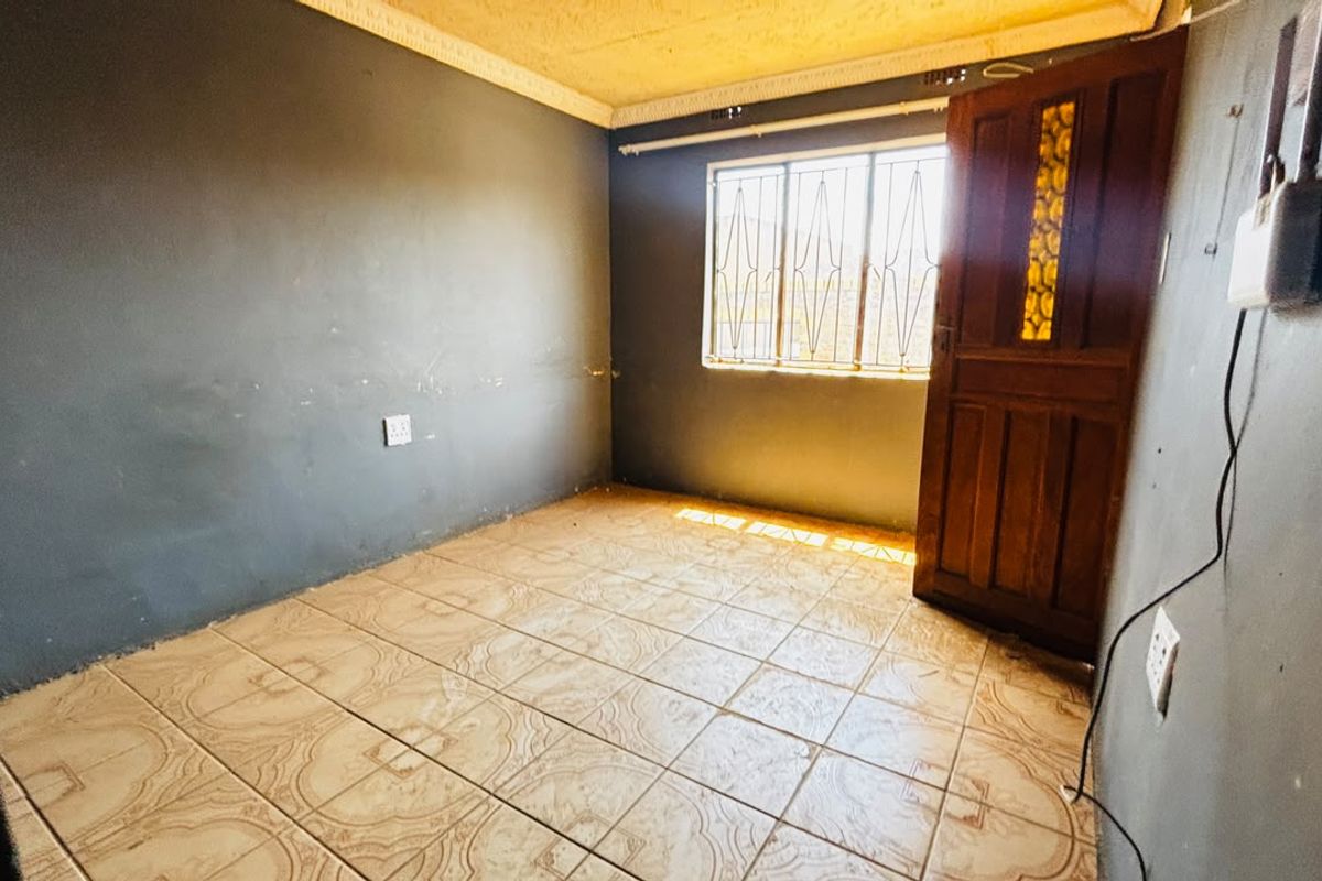 An empty bedroom featuring tiled flooring, security bars on the window, and a timber door, requiring cosmetic updates to the walls.