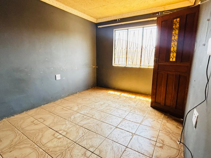 An empty bedroom featuring tiled flooring, security bars on the window, and a timber door, requiring cosmetic updates to the walls.