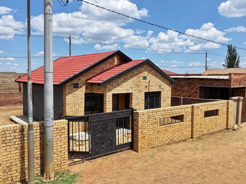 A single-story face brick house featuring a red tiled roof, enclosed by a perimeter brick wall with a metal gate.