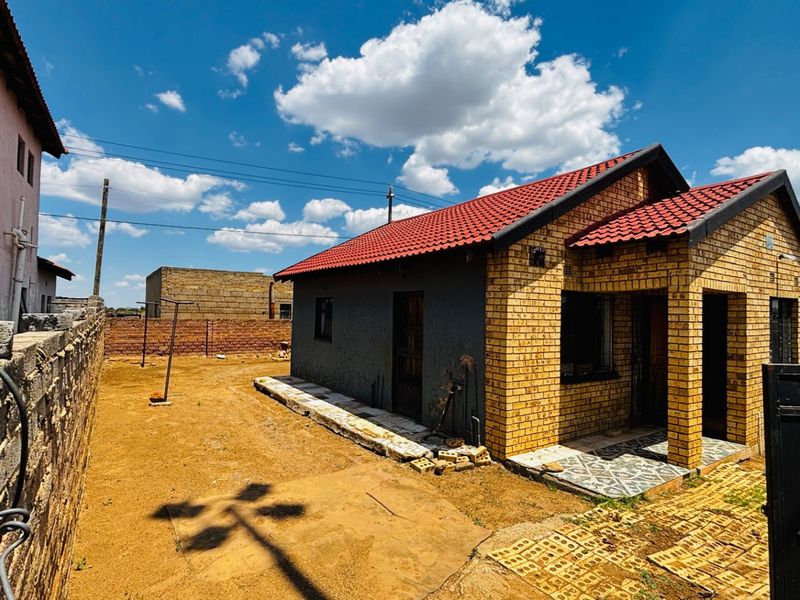 Front view of a face brick house featuring a red tiled roof and a covered entrance porch, set on a plot with paved areas.