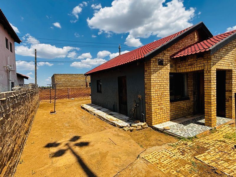 Front view of a face brick house featuring a tiled roof, covered entrance, and a large, undeveloped yard area enclosed by a perimeter wall.