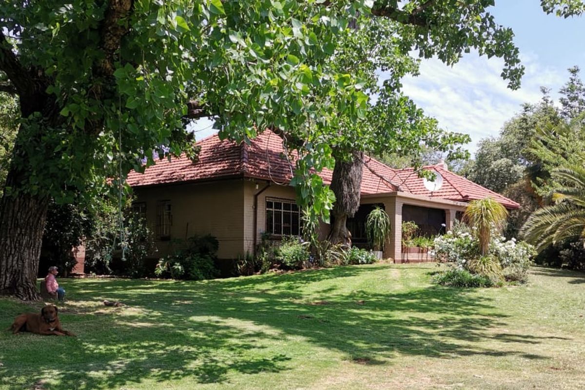 A view of the rear elevation of a brick farmhouse featuring a terracotta tile roof and a covered patio set within a mature garden.