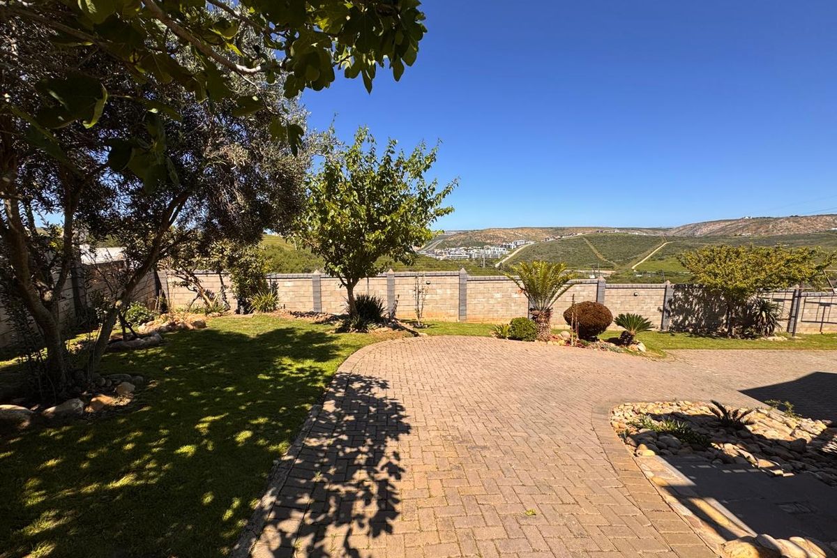 A well-maintained rear garden featuring a paved driveway, established trees, and a solid boundary wall with scenic hillside views.