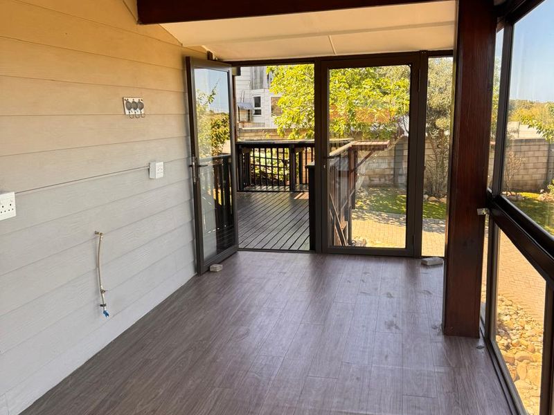 A bright, enclosed patio area featuring timber-look flooring, exposed structural beams, and large glass doors opening onto an outdoor deck.