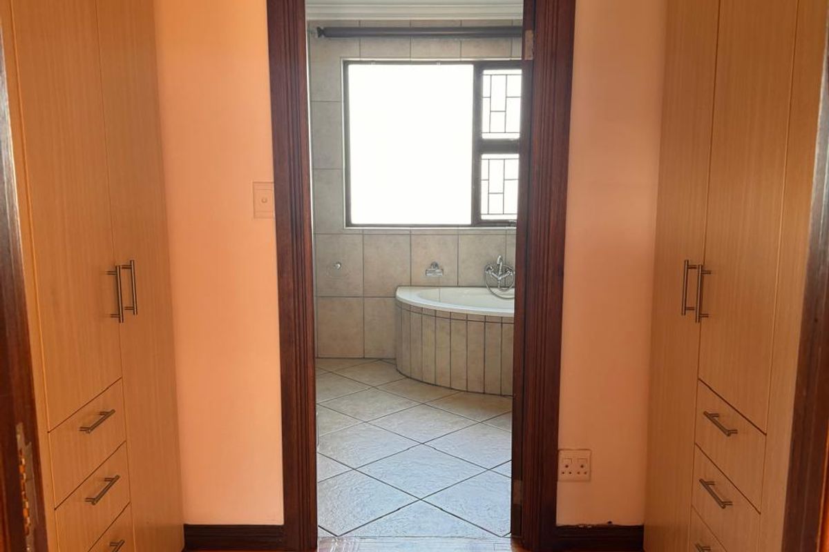 A walk-through dressing area featuring extensive built-in wooden cupboards and laminate flooring, leading directly into an en-suite bathroom with a corner bathtub.