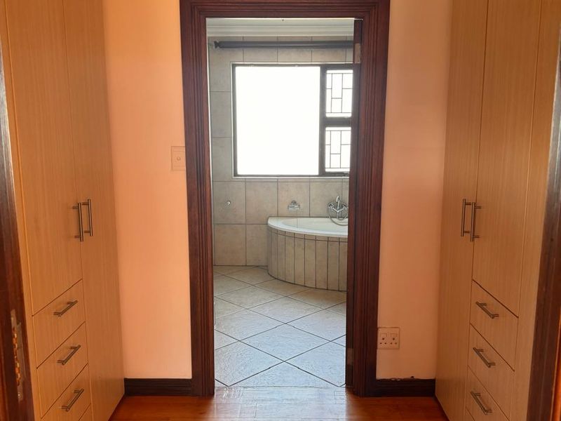 A walk-through dressing area featuring extensive built-in wooden cupboards and laminate flooring, leading directly into an en-suite bathroom with a corner bathtub.