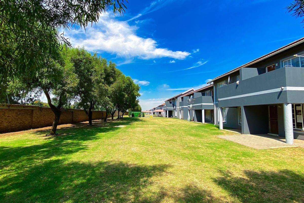 A view of the rear apartment complex facade featuring a well-maintained communal lawn, paved patio areas, and a brick boundary wall.