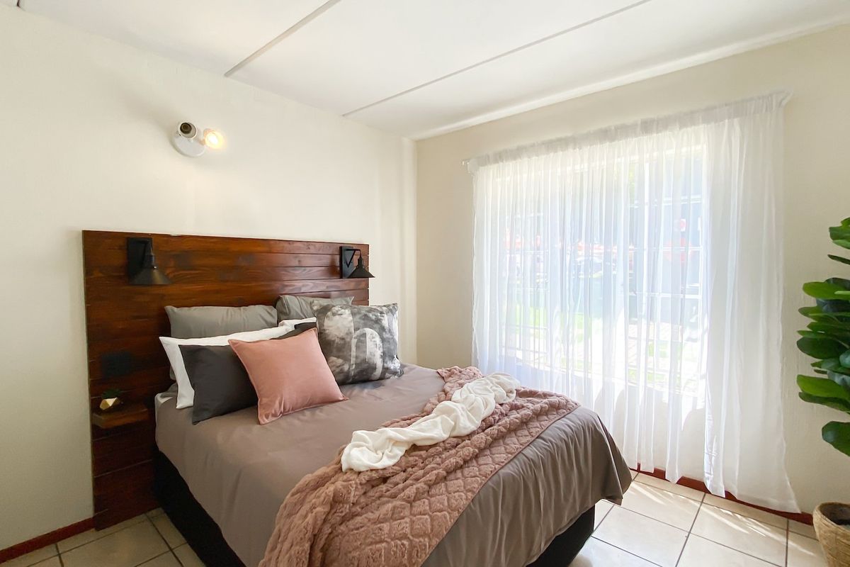A well-lit bedroom featuring a custom wooden headboard, wall-mounted lighting, and neutral tiled flooring with large window access.