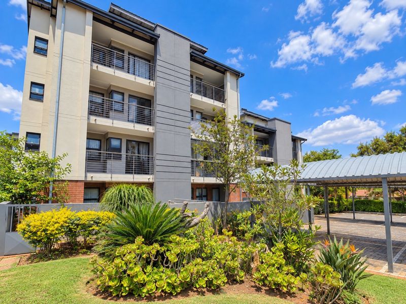 A modern apartment building exterior featuring private balconies, a well-maintained garden, and covered parking bays within a secure complex.