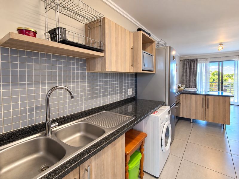 Modern open-plan kitchen featuring light wood cabinetry, granite countertops, tiled backsplash, and integrated appliances, flowing into a bright living area with balcony access.