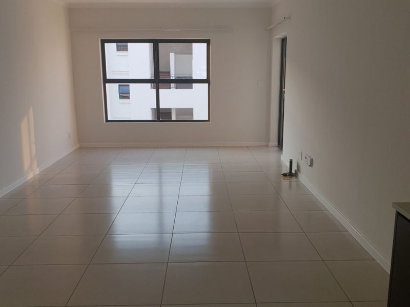 A bright, empty living area featuring neutral tiled flooring and large aluminium-framed windows, offering a clean and versatile space ready for furnishing.