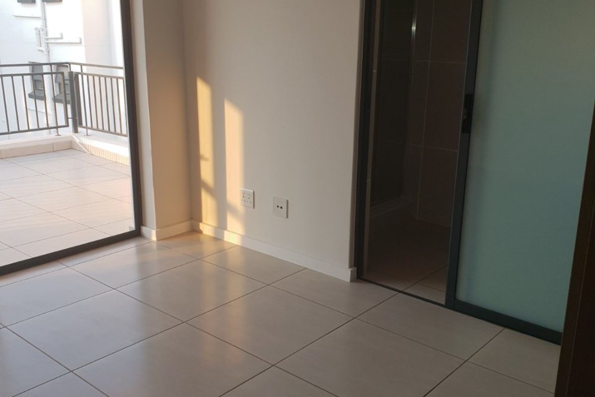 A bright, empty bedroom featuring neutral tiled flooring, a sliding door leading to a private balcony, and a frosted glass door accessing an en-suite bathroom.