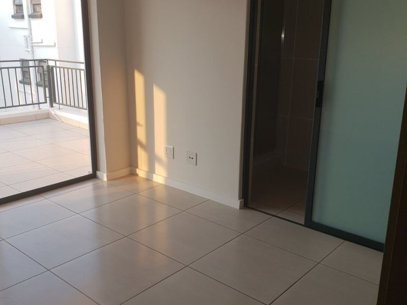 A bright, empty bedroom featuring neutral tiled flooring, a sliding door leading to a private balcony, and a frosted glass door accessing an en-suite bathroom.