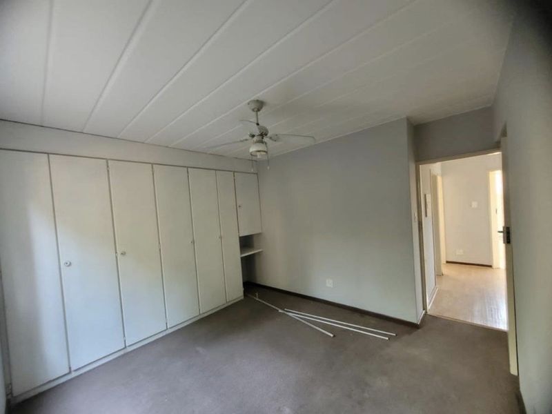 A vacant bedroom featuring extensive built-in cupboard storage, a ceiling fan, and a panelled ceiling, requiring cosmetic updates to the flooring and finishes.