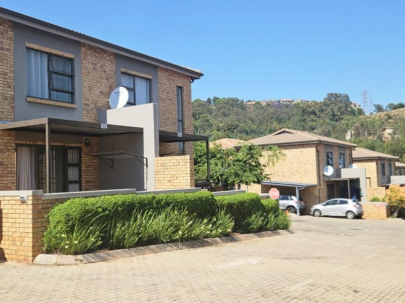 A view of the front facade of a face brick townhouse complex featuring paved driveways, covered patios, and established garden hedges.