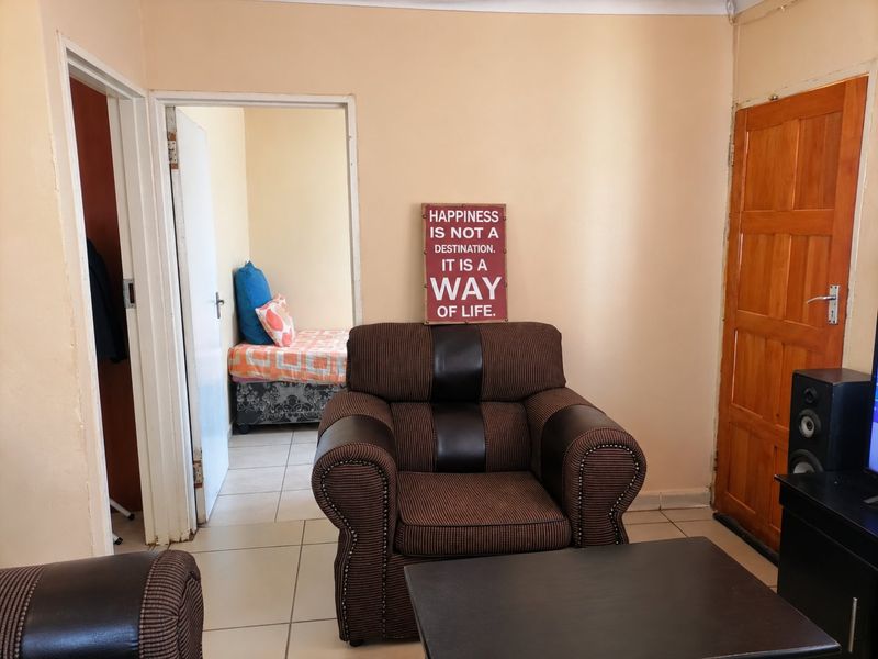A living room area featuring tiled flooring and a wooden door, with an adjacent view into a bedroom space.