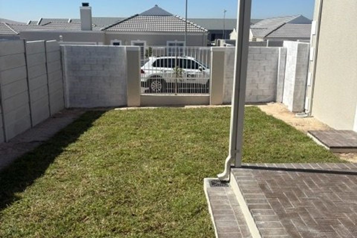 A neat rear garden featuring a paved patio area, a small lawn, and secure boundary walls with a metal gate.