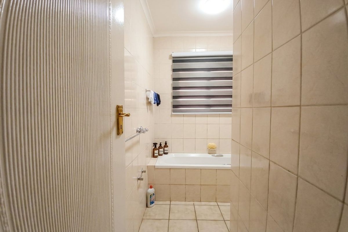 A functional bathroom featuring a built-in bathtub, neutral ceramic wall and floor tiling, and a window with a roller blind.