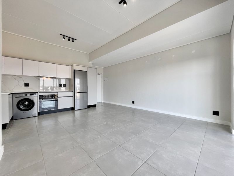 Modern open-plan living area featuring a sleek kitchen with built-in appliances, tiled flooring, and contemporary track lighting in a clean, neutral space.