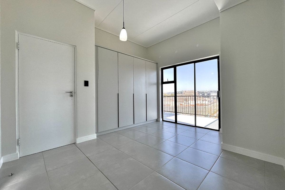 A bright, modern bedroom featuring neutral tiled flooring, ample built-in cupboard space, and a sliding glass door leading to an outdoor balcony.