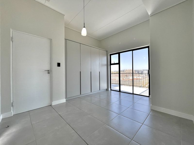 A bright, modern bedroom featuring neutral tiled flooring, ample built-in cupboard space, and a sliding glass door leading to an outdoor balcony.