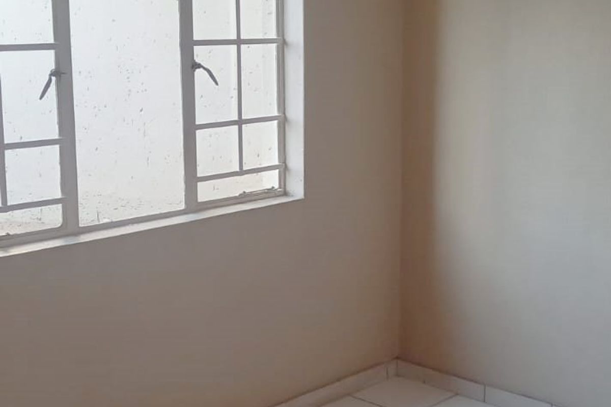 A clean, empty bedroom featuring white tiled flooring, neutral walls, and a window fitted with security bars.